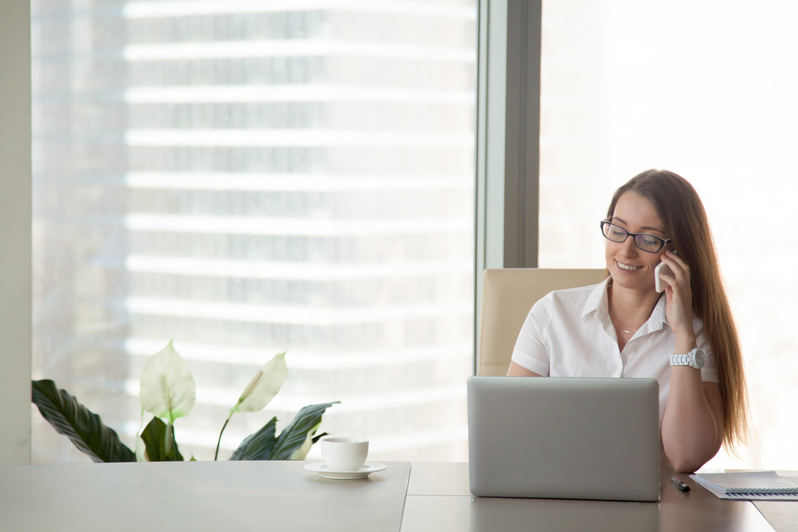 Young smiling businesswoman talking on the phone at workplace, happy office worker making business or personal call sitting at work desk, checking cell voicemail, corporate mobile communications